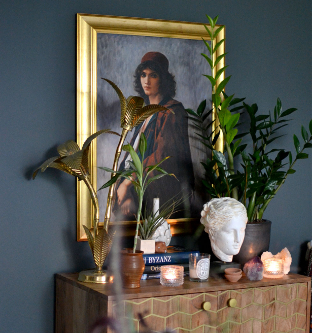 Young Bohemian Serb by Charles Landelle refined with a golden style frame. An elegant home decor setup featuring a classic oil painting with a gold frame, alongside a selection of indoor green plants, a marble bust, and decorative candles, all arranged on